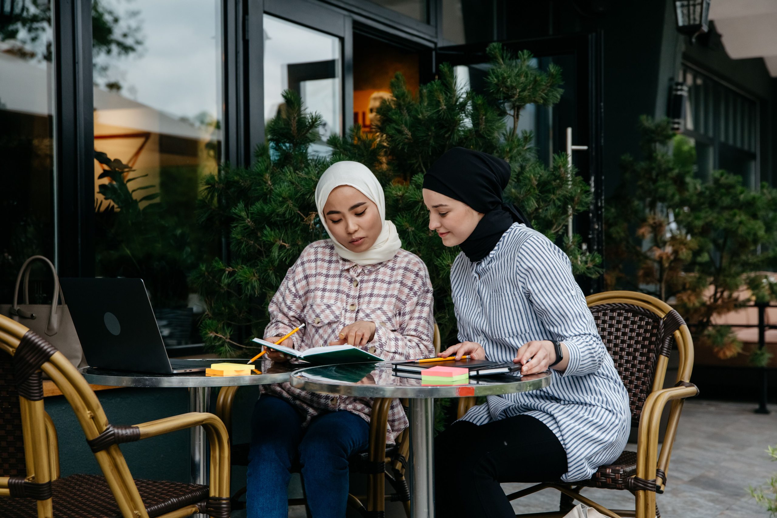 Two women in hijabs studying together at an outdoor cafe table.
